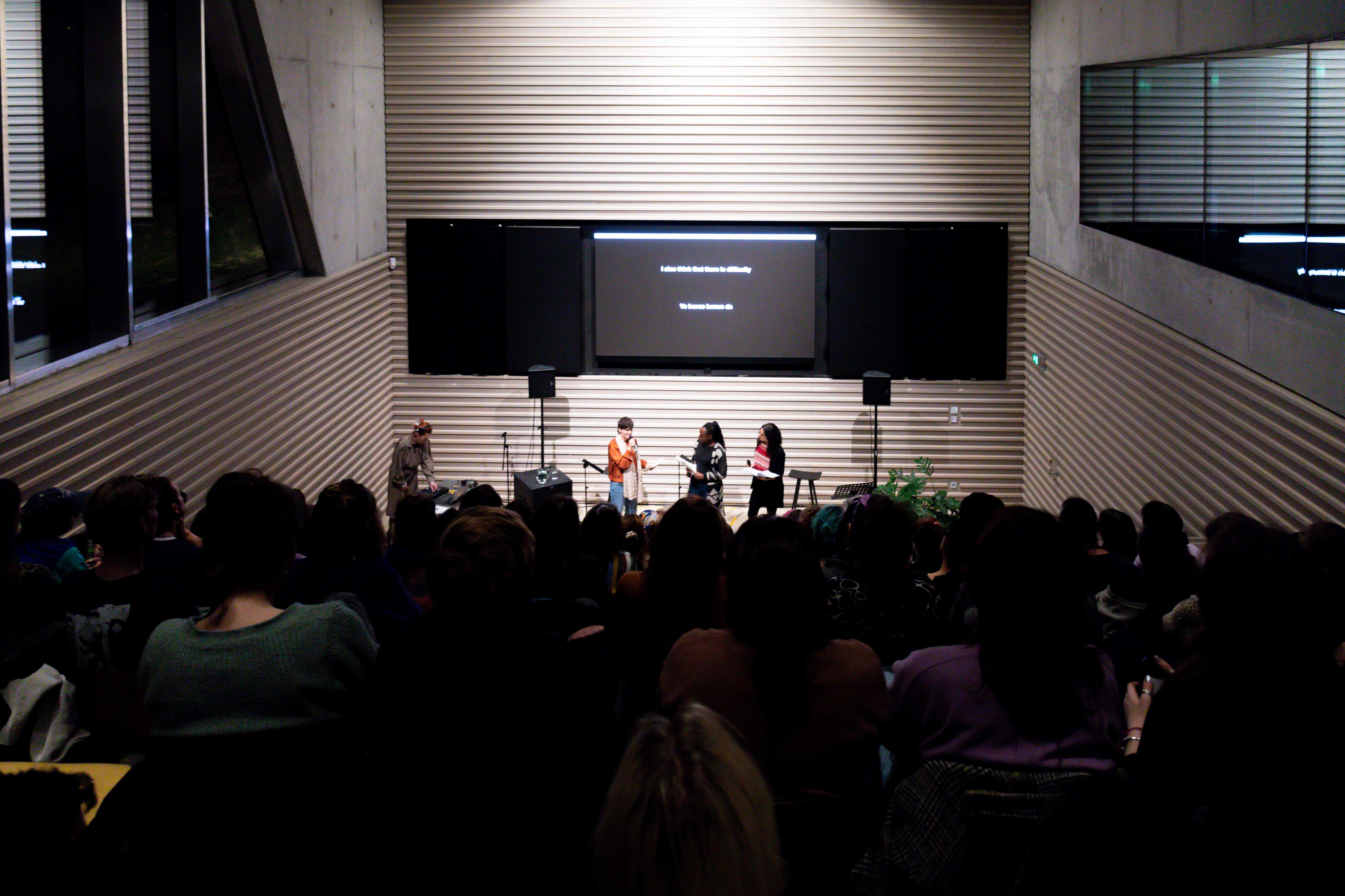 Auftrittssituation: Ein großer Raum mit einer herabsteigender Tribüne, auf der viele Menschen sitzen. Auf der Bühne, im Zentrum des Bildes befinden sich die vier Künstler*innen. Eine von ihnen spricht in das Mikrofon, die anderen schauen sie an. Über ihnen befindet sich ein Screen, auf dem die Untertitel des Stückes (weiße Schrift, schwarzer Hintergrund) zu sehen sind. 