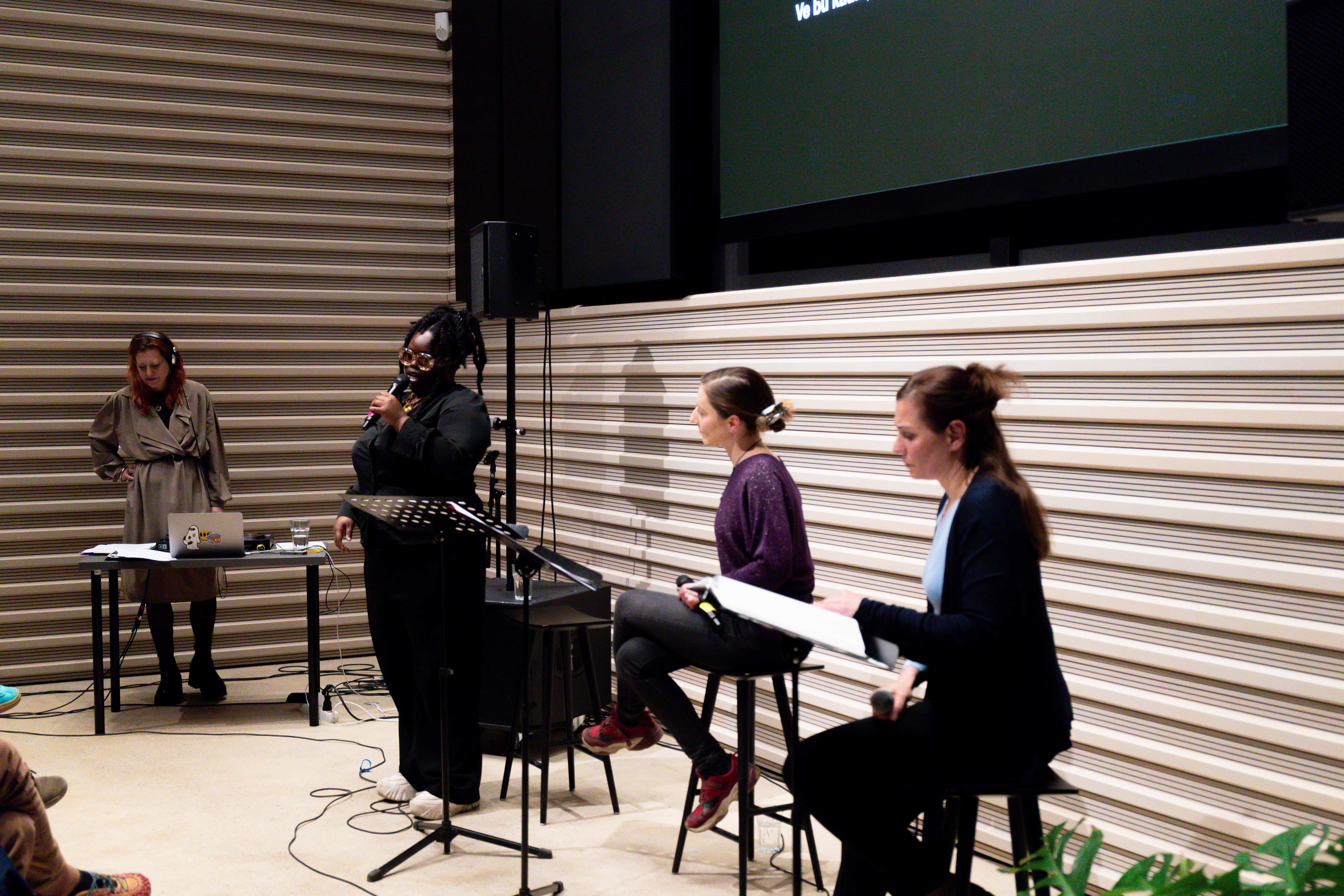 Eine Auftrittssituation: Vier Künstler*innen befinden sich auf der Bühne. Vor ihnen befinden sich Notenständer mit Textbüchern. Es handelt sich um drei Schauspieler*innen und eine DJ Person. Eine der Schauspieler*innen spricht in das Mikrofon.