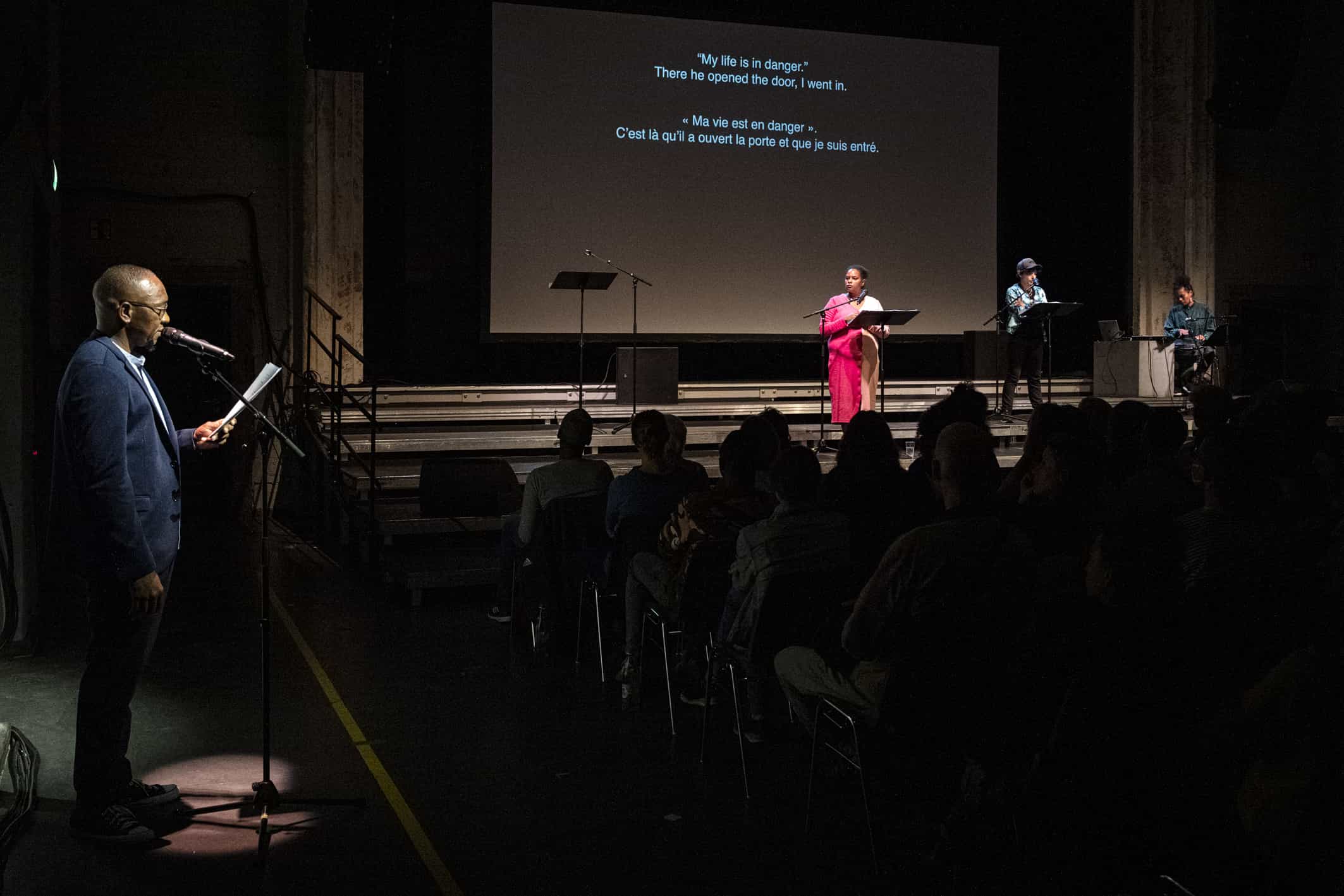 Auf dem Bild sind mehrere Menschen auf einer Bühne zu sehen. Links steht eine Person, die ins Mikrofon spricht. Sie steht auf einer anderen Bühne als die anderen Künstler*innen. Es befindet sich Publikum im Saal. Es ist eine große Leinwand zu sehen, auf der die Untertitel des Stückes (weiße Schrift, schwarzer Hintergrund) zu sehen sind. 